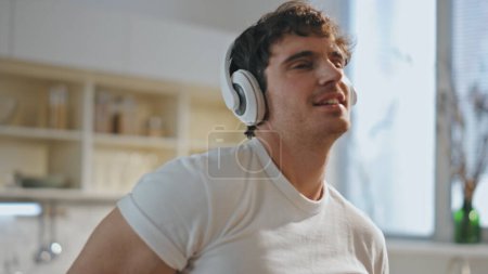 Cheerful man cooking in headphones dancing having fun at cozy kitchen close up. Happy smiling guy listening music in earphones preparing toast for breakfast. Handsome man singing favorite song at home