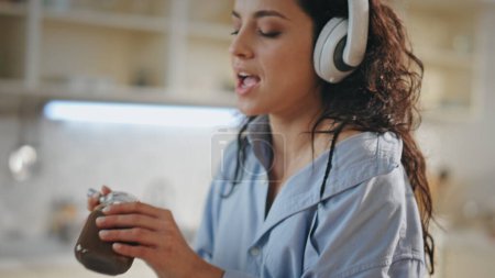 Cooking woman listening music in modern wireless headphones at kitchen closeup. Attractive young girl singing tasting chocolate paste from knife. Carefree relaxed housewife preparing food in earphones