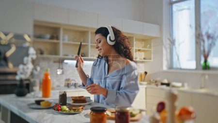 Singing woman preparing breakfast in headphones at comfortable home kitchen close up. Relaxed joyful girl listening favorite song in headset cooking alone. Attractive happy lady making tasty toasts.
