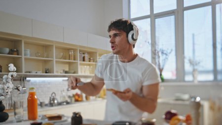Guy in earphones preparing food dancing along favorite song at home close up. Funny joyful man wearing wireless headset listening music cooking toast with chocolate pasta. Handsome singing at kitchen.