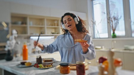 Girl in headphones cooking breakfast at modern kitchen. Carefree happy woman singing along favorite song in earphones preparing chocolate toasts at home. Carefree brunette dancing enjoying music.