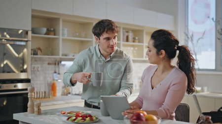 Photo for Married couple enjoying breakfast at modern kitchen together. Beautiful wife showing tablet computer to husband drinking morning coffee. Handsome man kissing woman looking watch hurrying on work. - Royalty Free Image