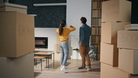 Couple watching at new house living room full cardboard boxes. Happy young family talking discussing interior rent apartment together. Married spouses planning home design enjoying purchased property.