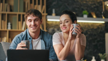 Smiling couple closing laptop finishing online meeting at home close up. Happy cheerful spouses saying goodbye video call sitting living room couch with coffee cup. Digital technology communication.