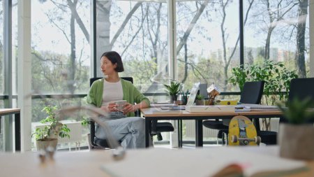 Entspannte Geschäftsfrau trinkt Kaffee und macht Pause im Büro. Ruhige asiatische Managerin, die sich am Computerarbeitsplatz ausruht. Lady sitzt in kreativem Coworking mit Panoramafenstern