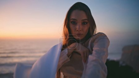 Closeup sensual woman moving hands smoothly with white fluttering fabric at sunset cliff. Young performer with magnetic gaze looking camera making graceful arms movements in front summer sky portrait