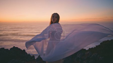 Sensual girl dancer waving fluttering white cloth performing contemporary dance in summer sunset. Romantic woman dancing with fabric on rocky hill at evening. Tender artist enjoying choreography 