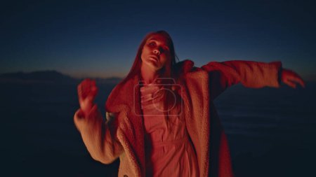 Relaxed girl contemporary dancer gesturing hands performing on ocean beach in neon light. Party chill woman wearing fur coat dancing modern style enjoying summer evening on seashore. Festive lifestyle