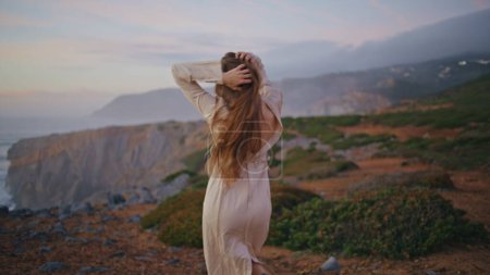 Carefree romantic lady tourist dancing on sunset sea cliffs zoom on. Relaxed serene woman enjoying stormy seascape at cloudy weekend closeup. Girl moving body seductively at cloudy beach wearing dress