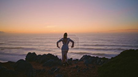 Sensual woman silhouette dancing sunset ocean landscape at summer. Beautiful dancer moving flexible body on rocky cliff. Seductive talented girl performing contemporary dance on evening hill back view