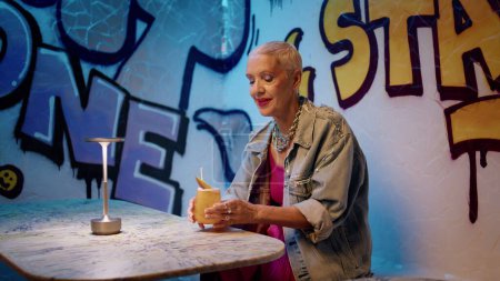Mode-Senior trinkt Cocktail-Stroh in Graffiti-Café. Fröhliche ältere Dame, die sich in einem Nachtclub ausruht und allein an einem alkoholischen Getränk nippt. Stylischer reifer Hipster genießt Wartetermin für den Ruhestand im trendigen Outfit