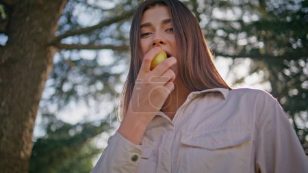 Woman eating apple park standing at sunlight closeup. Smiling beautiful lady delighting in taste of fresh fruit on sunny nature enjoy healthy snack outdoors. Happy girl chewing in front green foliage.