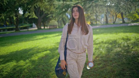 Relaxed girl walking in sunlight park exuding casual elegance. Happy woman enjoying serene nature at leisurely stroll holding water. Portrait showcases radiant female in peaceful outdoor recreation.