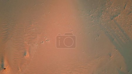 Sand-Fußabdrücke aus der Luft, die strukturierte Dünen bei Sonnenuntergang durchqueren. Die in orangefarbenes Dämmerlicht getauchte Landschaft bietet enorme Weiten für eine ruhige Erkundung. Naturmuster offenbaren ruhige, unberührte Wildnis