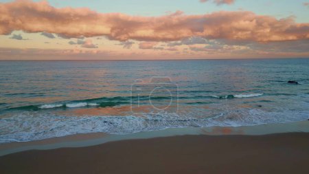 Luftaufnahme des Sonnenuntergangs an der Küste in rosa Farbtönen. Die Dämmerung senkt sich über den ruhigen Sandstrand, während die Wellen sanft die ruhige Küste umrunden. Weite Ozeane treffen heiteren Himmel in der Dämmerung spiegelt die Natur friedliche Schönheit.
