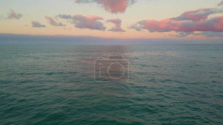 Drone soft ocean sunset bringing pink hue to clouds over calm tranquil ocean. Horizon stretching vast serene under evening twilight. Sea waters reflecting undisturbed dusk beauty. Marine panorama