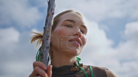 Relaxed girl warming sun at cloudy sky closeup. Smiling fashion model posing with stick on nature journey. Serene woman traveling in wilderness. Focused adventurer embodying spirit of independence.