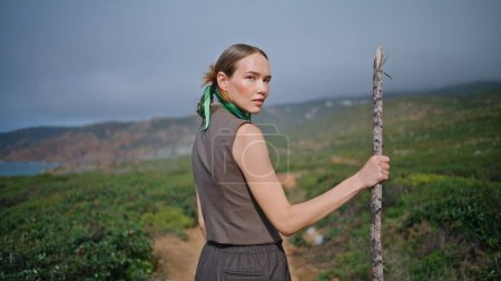 Outdoor woman going hill on summer day. Smiling hiker turning camera hold stick enjoying hiking adventure in wilderness. Calm girl walking path exploring green rugger terrain in sunlight rear view.