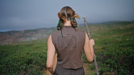 Woman journeying green hill with stick alone. Rear view serene girl walking path in solitude. Peaceful traveler admiring rugged beauty tranquil mountains landscape. Unknown lady wandering in sunlight