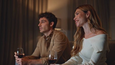 Couple spending first date in cozy evening apartment holding wine glasses close up. Smiling woman talking to handsome man watching tv. Cute pair shying spending evening weekend together. Love concept