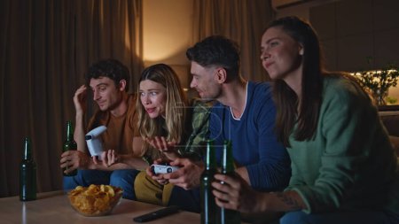 Freundliche Spieler, die abends zu Hause Videospiele spielen. Lächelnde Jugendliche, die Konsolen für Videospiele benutzen, um gemeinsam auf der Wohnzimmercouch zu sitzen. Fröhliche unbeschwerte Freunde genießen Unterhaltung am Wochenende in der Wohnung.