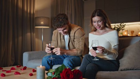 Cute young people dating in cozy apartment with candles. Smiling shying man hugging beautiful woman sitting home couch. Happy romantic couple holding wine glasses start watching tv in living room.