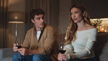Cute couple watching tv at romantic home atmosphere holding wine glasses. Shying man trying to flirt with beautiful woman sitting cozy sofa. Young pair spending first date in apartment with candles.