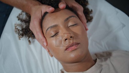 Téléchargez les photos : Canapé allongé patient ostéopathe avec vue de dessus les yeux fermés. Femme afro-américaine détendue rendant visite à un physiothérapeute dans une clinique de santé. Un homme méconnaissable masse la tête de la presse. Concept de soins physiques - en image libre de droit