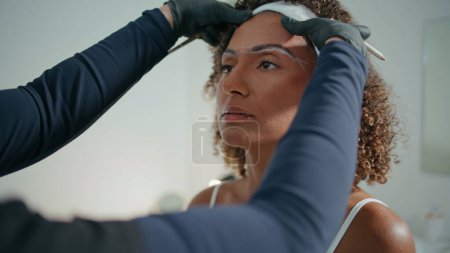 Cosmetologist shaping woman brows in beauty salon closeup. Curly african american girl visiting cosmetician for correction procedure in clinic. Mature lady consulting mark face with pencil in gloves