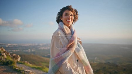 Femme élégante marche colline en manteau élégant le week-end soir. Joyeux voyageur souriant radieusement sur fond côtier pittoresque. Touriste branché mettant en valeur la mode et la confiance dans l'aventure pittoresque.