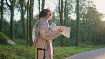 Explorer looking direction on map standing roadside at autumn forest. Beautiful stylish woman with suitcase choosing road in scenic woodland. Elegant female traveler exploring route in woods nature.