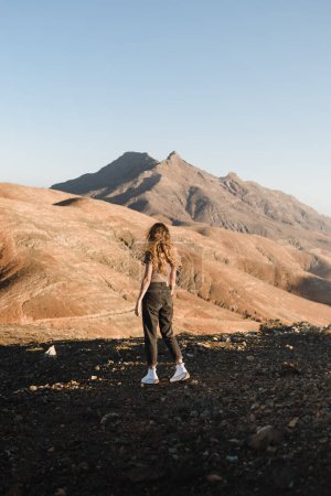 Foto de Mujer joven con pelo fluido y atuendo urbano caminando al atardecer en un paisaje desértico de las montañas de fuerteventura durante sus vacaciones en las islas canarias - Imagen libre de derechos