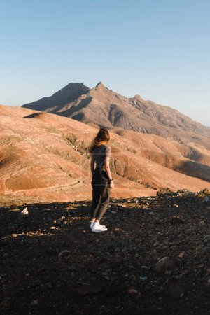 Téléchargez les photos : Jeune femme avec tenue urbaine debout regardant le coucher du soleil dans un paysage désertique des montagnes de fuerteventura pendant ses vacances dans les îles Canaries - en image libre de droit