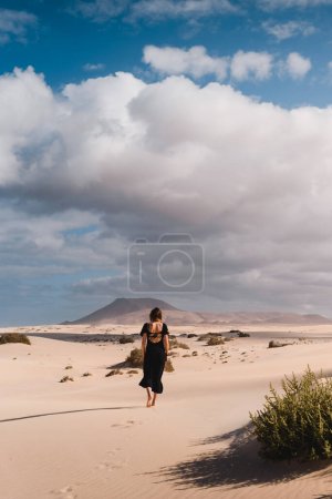 Téléchargez les photos : Mujer con vestido andando por el desierto de las dunas de corralejo en Fuerteventura, en verano, durante un viaje vacacional por las islas canarias. - en image libre de droit