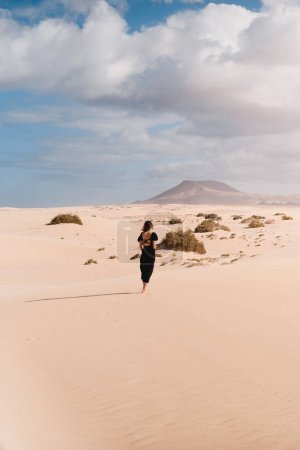 Téléchargez les photos : Femme en robe avec ses pieds regardant l'horizon dans les dunes de Corralejo à Fuerteventura, en été, lors d'un voyage de vacances à travers les îles Canaries. - en image libre de droit