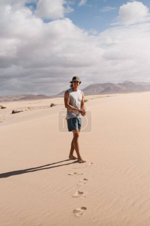 Téléchargez les photos : Heureux jeune homme marchant sur le sable désert des dunes de Corralejo à Fuerteventura, en été, lors d'un voyage de vacances à travers les îles Canaries - en image libre de droit