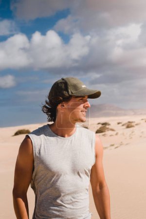 Téléchargez les photos : Portrait photo de jeune homme souriant avec bonnet dans le désert des dunes de Corralejo à Fuerteventura, regardant l'horizon, en été, lors d'un voyage de vacances à travers les îles Canaries. - en image libre de droit
