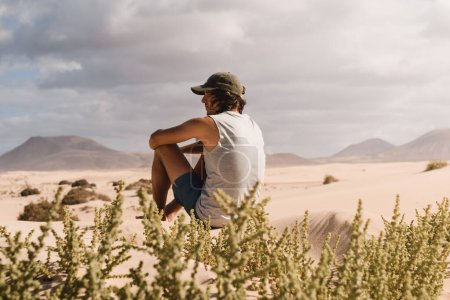 Téléchargez les photos : Jeune homme coiffé assis sur le sable désert des dunes de Corralejo à Fuerteventura, regardant l'horizon, en été, lors d'un voyage de vacances à travers les îles Canaries. - en image libre de droit
