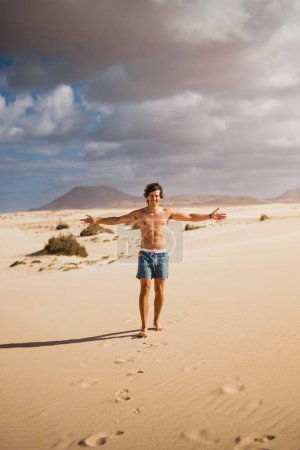 Téléchargez les photos : Jeune homme marchant heureux et torse nu sur le sable du désert des dunes de Corralejo à Fuerteventura, en été, lors d'un voyage de vacances à travers les îles Canaries. - en image libre de droit