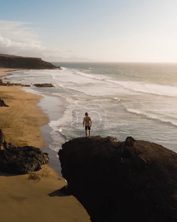 Téléchargez les photos : Jeune homme au coucher du soleil sur une falaise rocheuse à Fuerteventura lors de son voyage à travers les îles Canaries - en image libre de droit