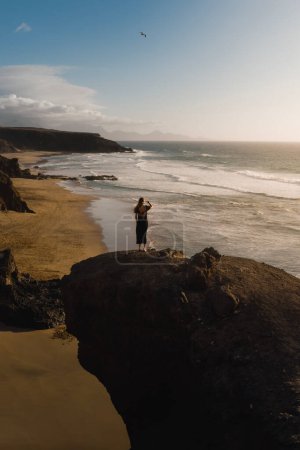 Foto de Joven disfruta de la puesta de sol sobre la playa en un acantilado de Fuerteventura durante su viaje por las Islas Canarias - Imagen libre de derechos