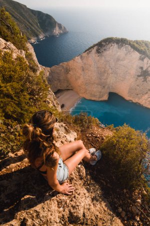 Foto de Mujer joven sentada en los acantilados sobre la playa de Navagio en la isla griega de Zakhyntos o Zante durante su viaje de verano - Imagen libre de derechos