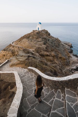 Téléchargez les photos : Fille marchant dans quelques escaliers sur son chemin vers une jolie église au sommet d'une falaise sur l'île grecque de Sifnos - en image libre de droit