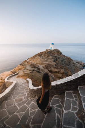 Foto de Mujer joven bajando unas escaleras en el camino a una iglesia en la parte superior de una roca junto al acantilado en la isla de Sifnos - Imagen libre de derechos