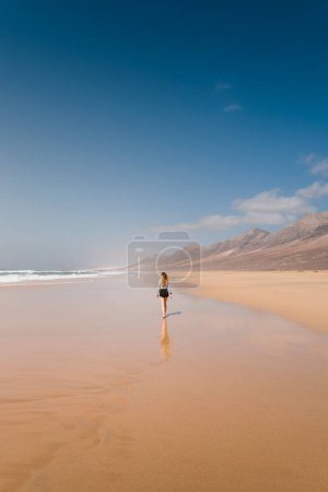 Téléchargez les photos : Jeune femme marchant au coucher du soleil le long du sable de la plage sauvage de cofete avec un paysage montagneux en arrière-plan. à fuerteventura. Pays-Bas. - en image libre de droit