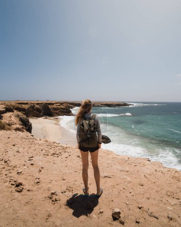 Foto de Joven mochilera mirando al mar desde los desiertos acantilados de la costa salvaje de la isla canaria de Fuerteventura durante su viaje de exploración - Imagen libre de derechos