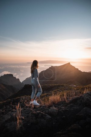 Foto de Mujer joven con atuendo urbano mirando una hermosa puesta de sol desde la cima de Teno en la isla canaria de Tenerife durante sus vacaciones turísticas - Imagen libre de derechos