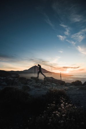Téléchargez les photos : Rétroéclairage d'un jeune homme profitant parmi les rochers de la montagne au coucher du soleil avec le volcan Teide en arrière-plan. Au cours de son voyage d'aventure à travers les îles Canaries de Ténérife - en image libre de droit