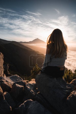 Jeune femme aux cheveux longs assise sur la montagne regardant le sommet du Teide lors de son voyage touristique à travers Tenerife, dans les îles Canaries