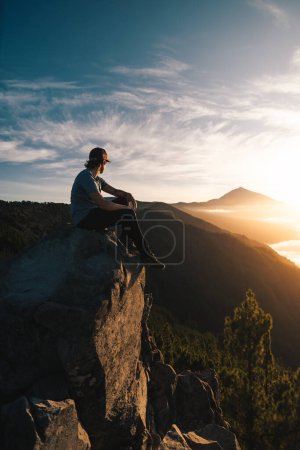 Photo for Young man with cap sitting on a rock observing the Teide peak at sunset during his touristic travel through the Canary Island of Tenerife - Royalty Free Image